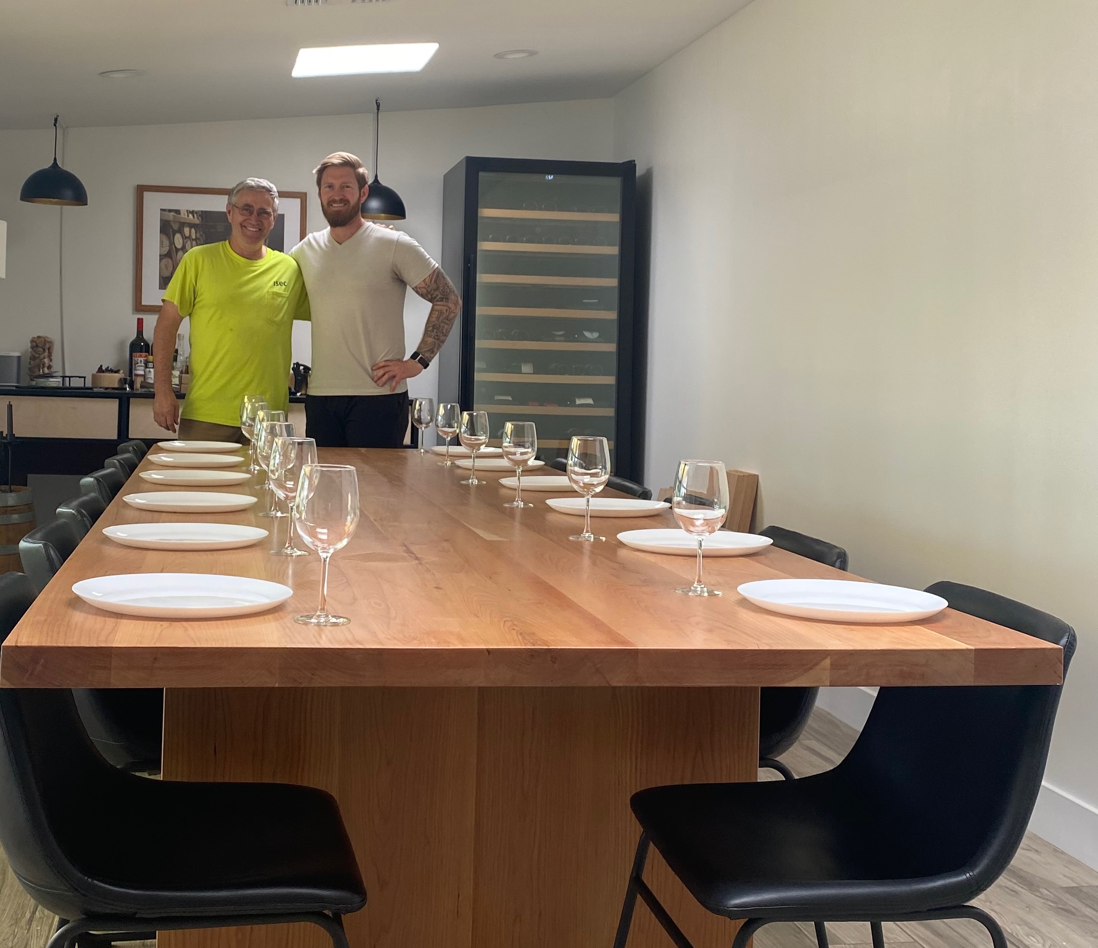 Josh and his father standing behind a long solid cherry dining table they built, set with plates and wine glasses